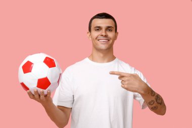 Yakışıklı genç adam pembe arka planda futbol topuna işaret ediyor.