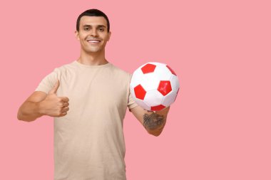 Elinde futbol topu olan yakışıklı genç adam pembe arka planda baş parmak hareketi yapıyor.