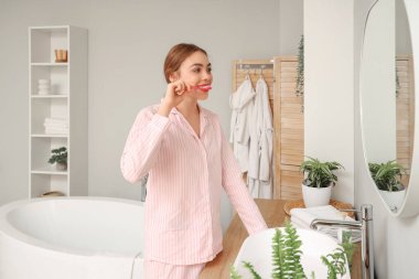 Güzel genç bir kadın banyoda aynanın yanında dişlerini fırçalıyor.