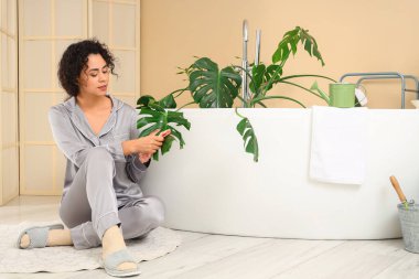 Genç Afro-Amerikalı kadın banyoda Monstera bitkisinin yapraklarını siliyor.
