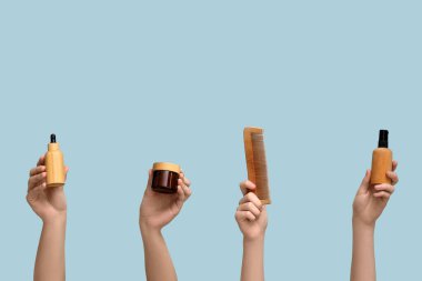 Farklı kozmetik ürünlere sahip kadın elleri ve mavi arka planda saç tarağı