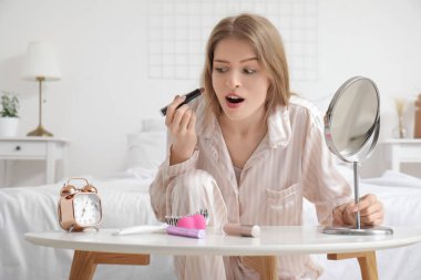Evdeki çalar saati kontrol ederken acele makyaj yapan genç bir kadın.