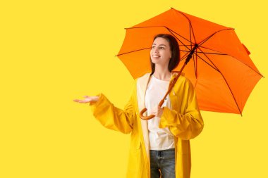 Sarı arka planda açık şemsiyesi olan yağmurluk giyen mutlu genç bir kadın.