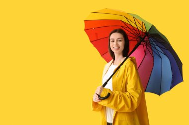 Sarı arka planda açık renkli şemsiyesi olan yağmurluk giyen mutlu genç bir kadın.