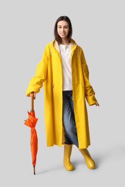 Gri arka planda şemsiyesi olan yağmurluk giyen mutlu genç bir kadın.