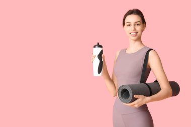Pembe arka planda bir şişe su ve yoga minderiyle güzel, genç ve mutlu bir kadın.