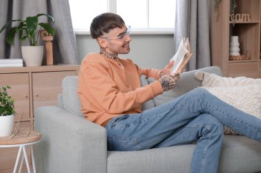 Gözlüklü genç adam evdeki kanepede kitap okuyor.