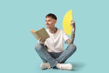 Elinde yelpaze olan dövmeli genç bir adam mavi arka planda kitap okuyor.