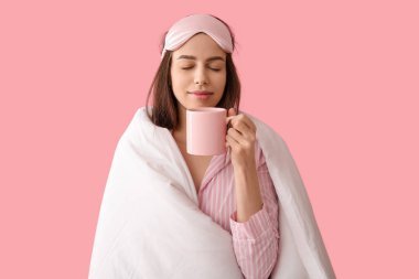 Pembe arka planda fincanı olan pijamalı ve battaniyeli genç bir kadın.