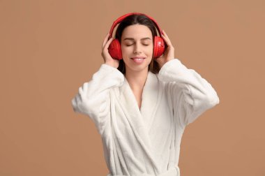 Bornozlu ve kulaklıklı genç bir kadın bej arka planda müzik dinliyor.