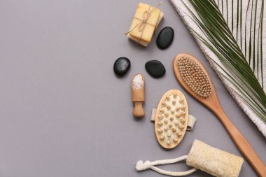 Masaj vücut fırçaları, spa taşları, sabun kalıpları, bir kepçe deniz tuzu ve gri arka planda palmiye yaprağı içeren kompozisyon