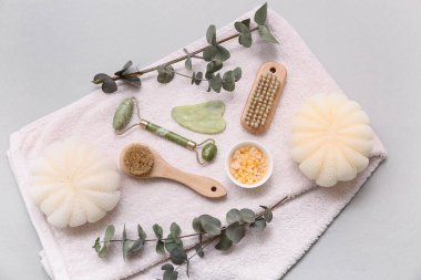 Yüz masajı aletleri, banyo süngeri, havlu, deniz tuzu, vücut masajı fırçaları ve beyaz arka planda okaliptüs dalları.