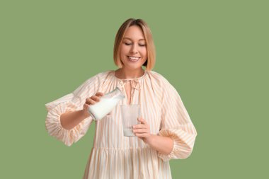 Güzel genç bir kadın yeşil arka planda şişeden bardağa taze süt döküyor.