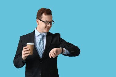 Gözlüklü genç adam elinde bir fincan kahveyle mavi arka plandaki saatlere bakıyor.