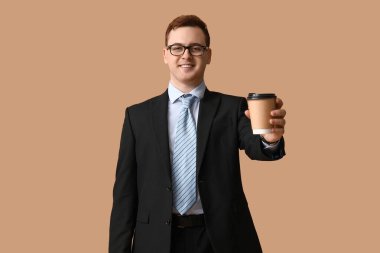 Takım elbiseli ve gözlüklü genç adam kahverengi arka planda bir fincan kahve tutuyor.