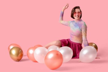 Pembe arka planda balonları olan şık giyinmiş mutlu, genç, modaya uygun bir kadın.