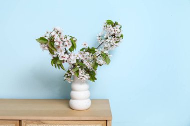 Mavi duvarın yanındaki masada çiçek açan dalları olan bir vazo.