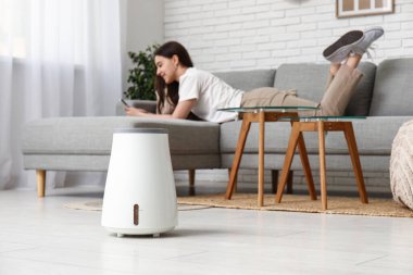 Moda oturma odasındaki modern nemlendiricinin yanındaki rahat koltukta cep telefonu kullanan genç bir kadın.