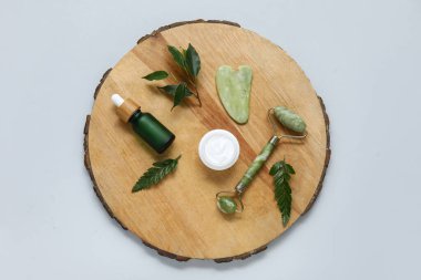 Tahta tahta silindir, serum, gua sha, kozmetik krem ve gri arka planda yeşil yapraklar.