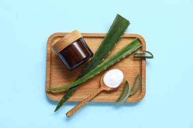 Ahşap tepsi, taze aloe yaprakları, kavanoz ve kepçe mavi arka planda kozmetik ürünü.