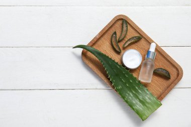Aloe vera yaprağı, ahşap tepsi, kavanoz ve beyaz ahşap arka planda bir şişe kozmetik ürünü.