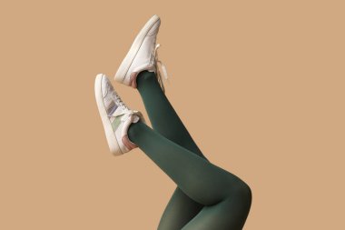 Yeşil taytlı genç kadın bacakları ve bej arka planda beyaz spor ayakkabılar.