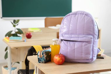 Sınıftaki masada elma suyu, meyve suyu ve kalem kutusuyla leylak rengi sırt çantası.