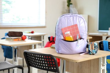 Sınıftaki masada meyve suyu ve kalem çantalı leylak rengi sırt çantası.