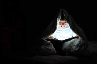 Karanlık yatak odasında battaniyenin altında el feneri okuyan küçük Afro-Amerikalı çocuk.