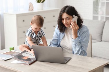 Evde cep telefonuyla konuşurken dizüstü bilgisayarla çalışan tatlı bir bebek ve genç anne.