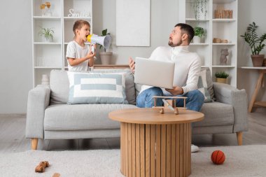 Küçük oğlu megafona bağırıyor ve şok olmuş babası evdeki kanepede dizüstü bilgisayarla çalışıyor.