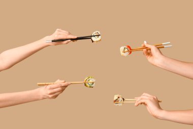 Kadın elleri, bej arka planda farklı lezzetli suşi ruloları olan çubukları tutuyor.