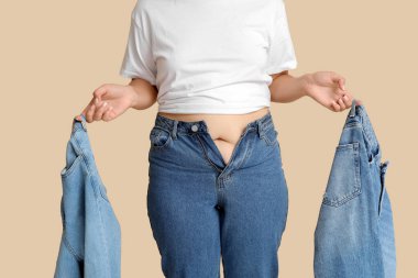 Bej arka planda farklı dar kot pantolonları olan şişman bir kadın. Aşırı ağırlık kavramı