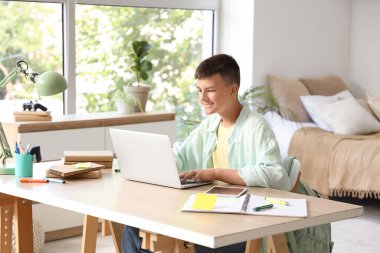 Teenage boy studying with laptop at table in bedroom
