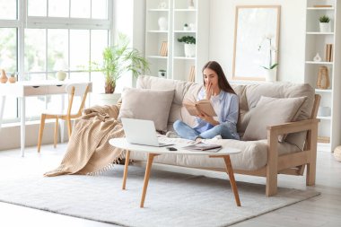 Evdeki kanepede dizüstü bilgisayarla kitap okuyan genç ve güzel bir kadın.