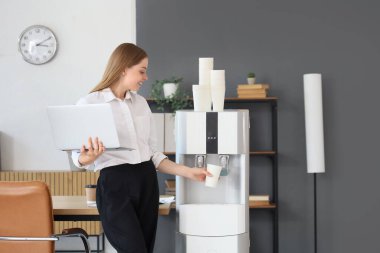 Dizüstü bilgisayarlı iş kadını ofisteki soğutucudan su dolduruyor.