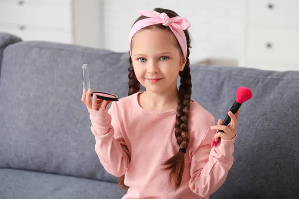 Cute little girl with blush and makeup brush sitting on sofa in living room