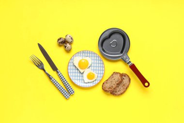 Kızartma tavalı, kalp şeklinde yumurta tabağı, bıldırcın yumurtası ve sarı arka planda çatal bıçak takımı.