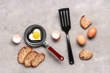 Composition with heart-shaped fried egg in frying pan, eggs, eggshell, spatula and bread on grunge background clipart