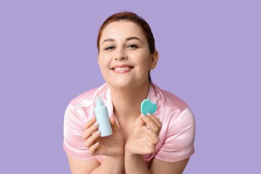 Elinde kalp şeklinde makyaj süngeri ve eflatun arkaplanda bir şişe kozmetik ürünü tutan güzel bir kadın.