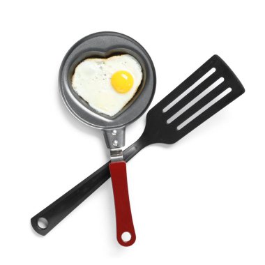 Beyaz arka planda kalp şeklinde sahanda yumurta ve spatula ile kızartma tavası.