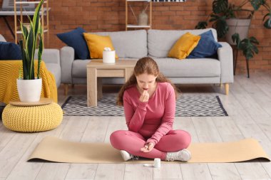 Hasta, boğazı ağrıyan ve evde yoga minderinde otururken ilaç alan genç bir kadın.