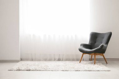 Odadaki ışık perdesinin yanındaki rahat koltuk ve halı.