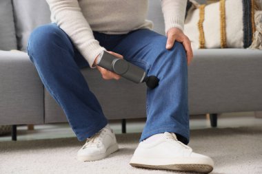 Olgun bir adam evdeki koltukta perküsyon masajıyla bacağına masaj yapıyor.