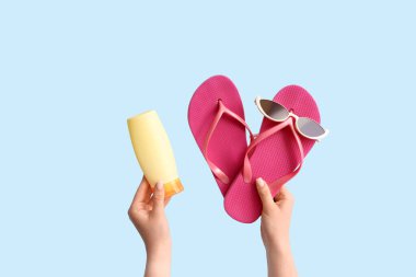 Mavi arka planda güneş gözlüklü, parmak arası terlikli ve güneş kremli bir kadın eli.