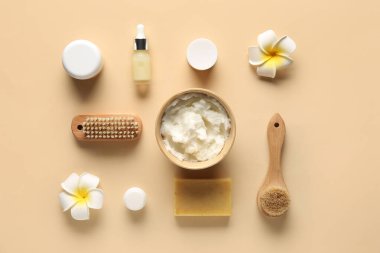 Bir kase tereyağı, bir şişe kozmetik yağı, sabun barı, tesisat çiçekleri ve bej arka planda masaj fırçaları.