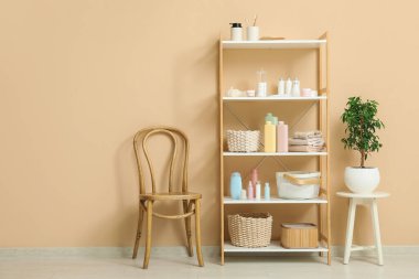 Bej duvarın yanında kozmetik ürünleri, sandalye ve bitki dolu raflar.