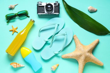Havalı parmak arası terlikler, bir şişe meyve suyu, fotoğraf makinesi ve turkuaz arka planda plaj aksesuarları.
