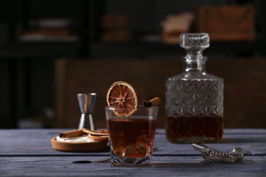 Bir bardak eski moda kokteyl, tarçın çubuğu, kurutulmuş portakal dilimi ve gri ahşap masada süzgeç.