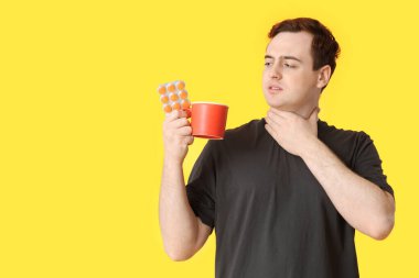 Boğazı ağrıyan genç adam, haplar ve sarı arka planda sıcak çay.